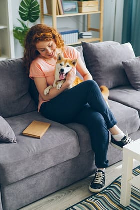 A woman with red hair embraces a dog while sitting on a gray couch in a cozy living room.